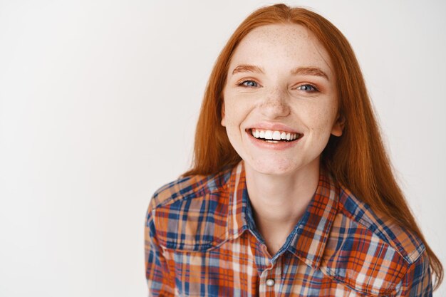 Close da bela modelo feminina ruiva com pele pálida e dentes brancos, sorrindo feliz na frente, em pé sobre uma parede branca