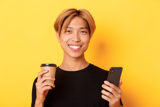 Close de um belo jovem asiático sorrindo feliz, usando smartphone e bebendo café para viagem, em pé sobre a parede amarela