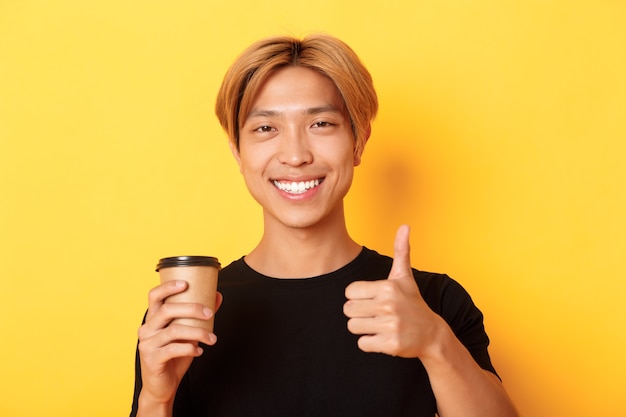Close de um cara bonito asiático satisfeito, recomendando café, segurando uma xícara de café e mostrando o polegar para cima em aprovação, sorrindo satisfeito com a parede amarela