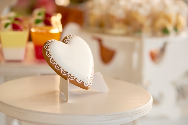 Close de um delicioso biscoito vitrificado em forma de coração em um suporte de madeira perto do candybar com diferentes sobremesas, como cupcakes amarelos e geléias vermelhas.
