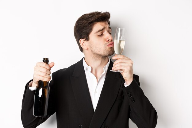 Close de um homem bonito de terno, beijando taça com champanhe, embebedando-se em uma festa, em pé contra um fundo branco