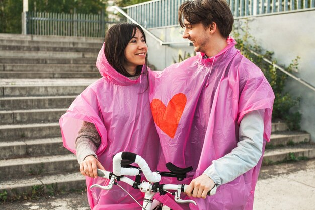 Close de um homem e uma mulher compartilhando uma capa de chuva de plástico rosa com um coração vermelho no centro