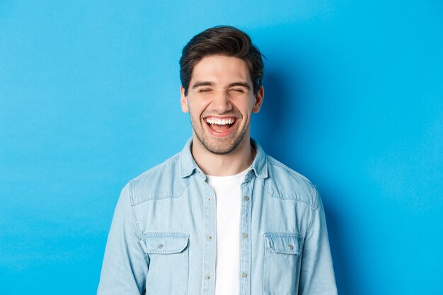 Close de um jovem barbudo feliz em roupas casuais, rindo e se sentindo alegre, em pé sobre um fundo azul