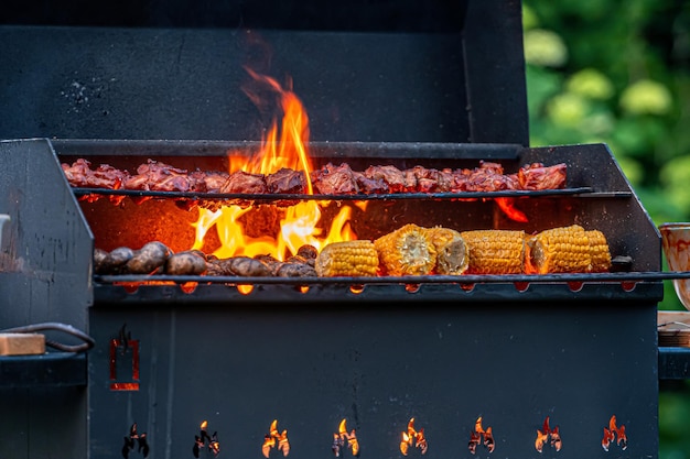 Close de uma grelha cheia de comida deliciosa, milho e carne assados em uma churrasqueira de jardim com chamas de fogo
