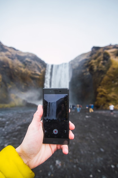 Close de uma mão gravando um vídeo da cachoeira skogafoss com um telefone na islândia
