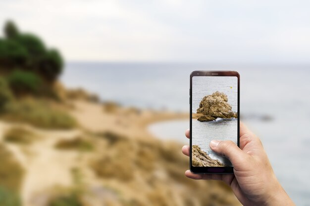 Close de uma mão segurando um telefone, tirando uma foto de uma praia