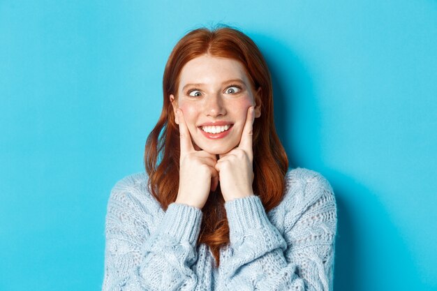 Close de uma menina adolescente ruiva engraçada fazendo caretas, apertando os olhos e apertando as bochechas, em pé contra um fundo azul