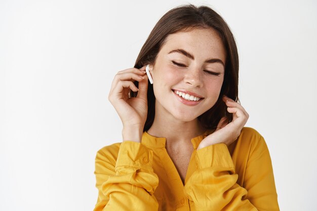 Close de uma mulher atraente, ouvindo música em fones de ouvido, sorrindo enquanto aprecia uma música arrepiante, em pé sobre uma parede branca