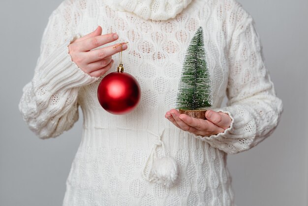 Close de uma mulher branca segurando uma pequena árvore de Natal em uma e uma bola vermelha na outra