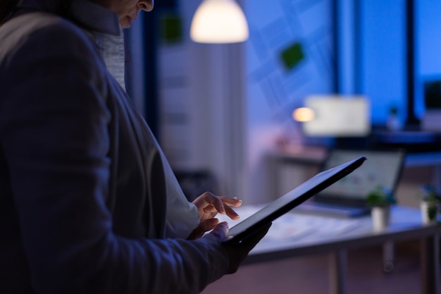 Close de uma mulher com as mãos digitando em um tablet, verificando os gráficos financeiros em pé no escritório de start-up tarde da noite