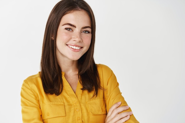 Close de uma mulher confiante e feliz, sorrindo, com os braços cruzados no peito como uma profissional, em pé sobre uma parede branca