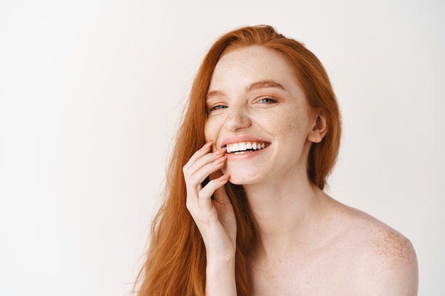 Close de uma mulher ruiva feliz com pele pálida perfeita, rindo e mostrando os dentes brancos, nua na parede do estúdio