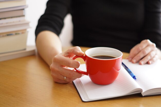 Close de uma mulher trabalhando ou estudando em casa com um café vermelho na mão