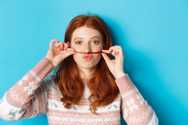 Foto grátis close de uma ruiva boba e engraçada fazendo bigode com uma mecha de cabelo e lábios franzidos, fazendo uma careta contra um fundo azul