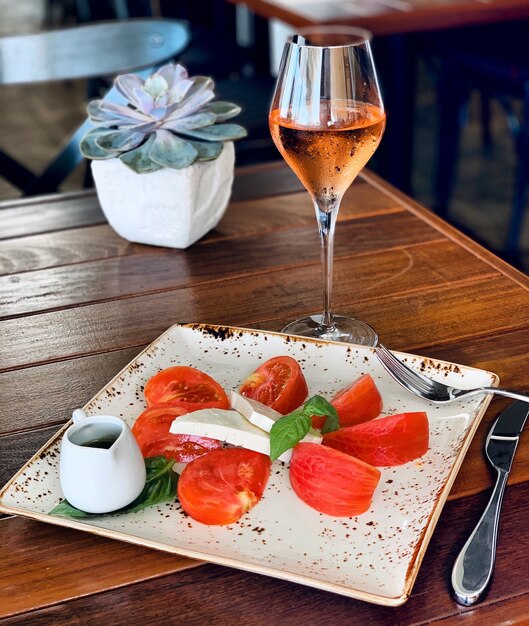 Close de uma salada com queijo e tomate em um prato quadrado com bebida alcoólica perto de uma planta