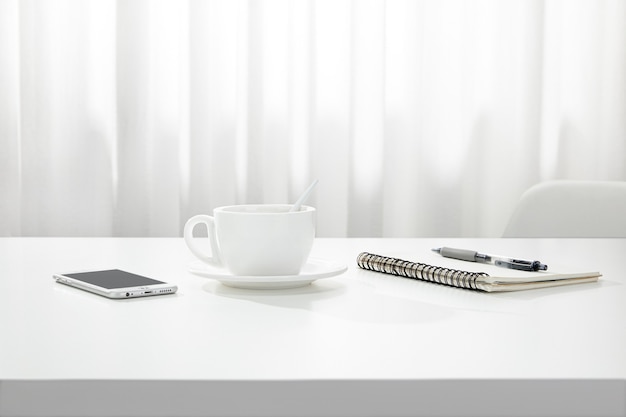Close de uma xícara de café, um caderno e uma caneta e um smartphone em uma mesa branca, dentro de casa