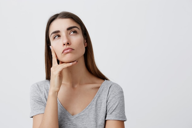 Close-up da bela encantadora jovem aluna caucasiana com cabelos longos escuros na elegante camiseta lisa, segurando o dedo perto do rosto, olhando de lado com expressão pensativa e relaxada