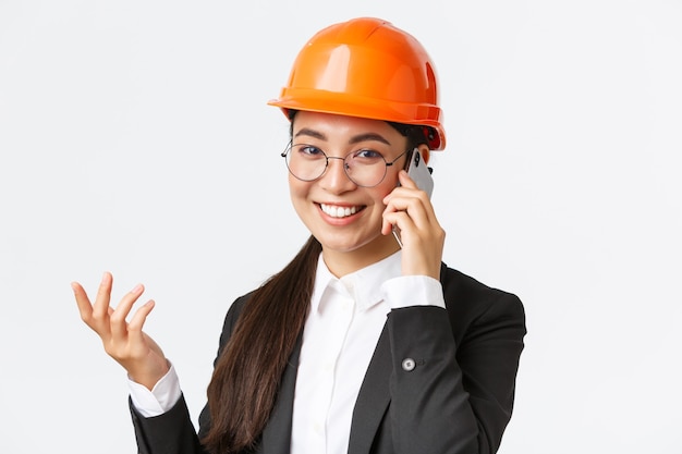 Close-up da empresária asiática gerencia a empresa, engenheiro com capacete de segurança e terno, conversando ao telefone, ligando para investidores, sorrindo enquanto fala sobre o smartphone, fundo branco