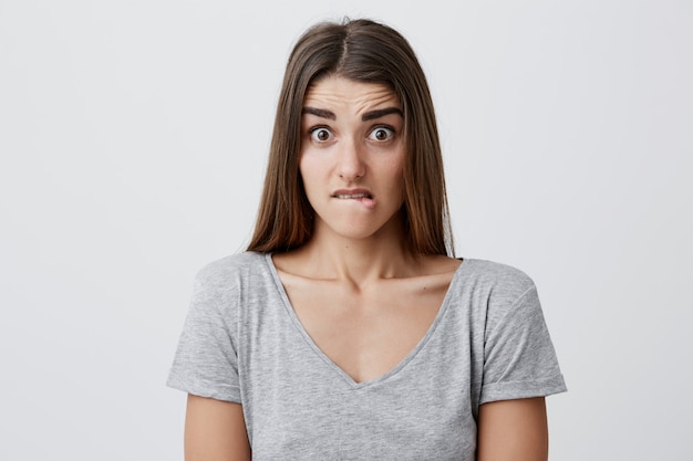 Foto grátis close-up da jovem atraente caucasiana aluna com longos cabelos escuros na camiseta cinza, olhando com expressão chocada e assustada, mordendo os lábios
