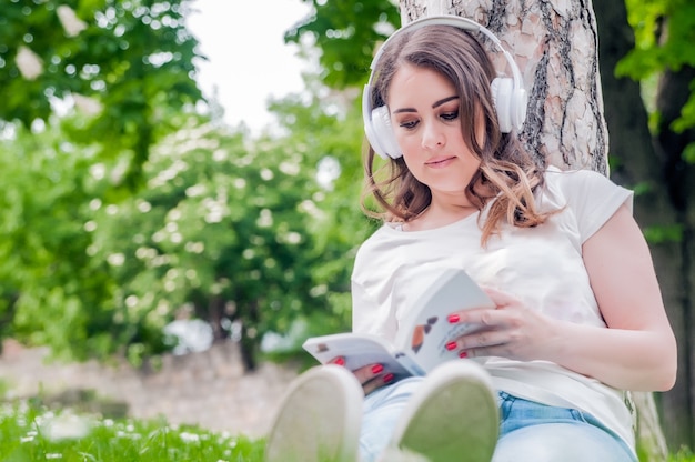 Close-up da jovem e relaxada mulher lendo e ouvindo música em fones de ouvido no dia da primavera. Conceito de tempo livre com mulher bonita relaxando ao ar livre com música, livros e café