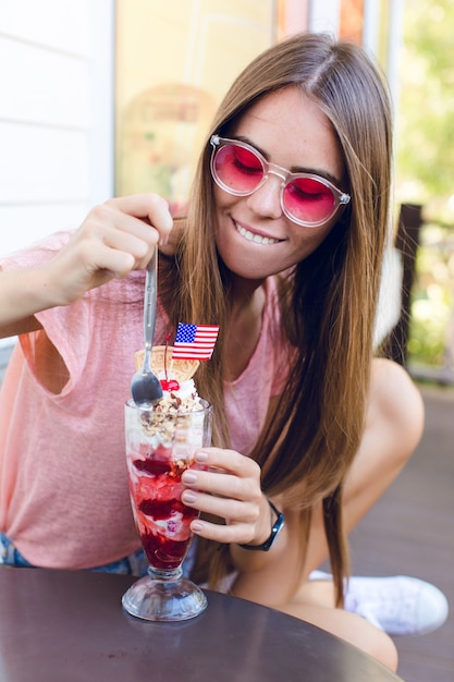 Close-up da linda garota sentada em uma cadeira tomando sorvete com cereja no topo com uma colher. Ela usa shorts jeans, blusa rosa e sorrisos. Ela tem óculos rosa