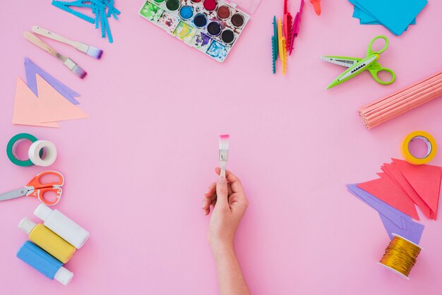 Close-up da mão de uma mulher segurando o pincel com a paleta de cores de água; pincel; papel; tesoura no pano de fundo rosa