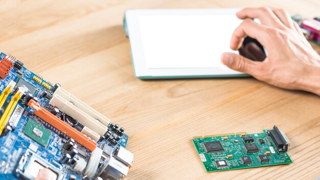 Close-up da mão tocando tablet digital com placa-mãe e circuito na mesa de madeira