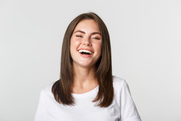 Close-up da menina morena feliz em t-shirt branca, rindo e sorrindo despreocupada para a câmera.