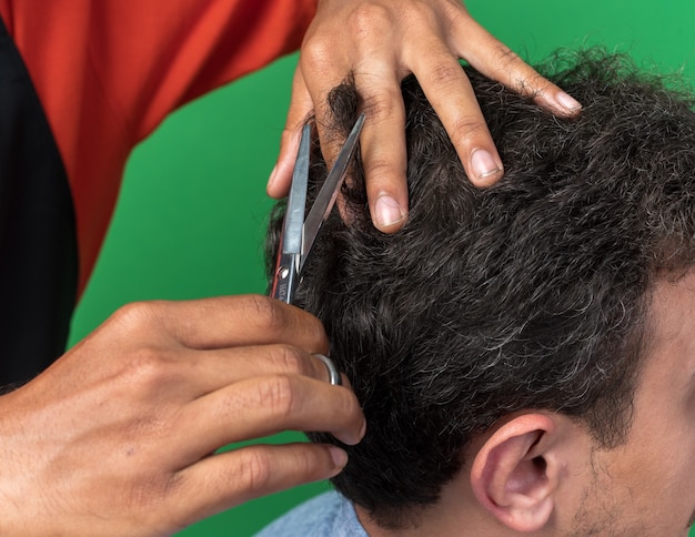 Close-up das mãos do barbeiro masculino cortando o cabelo de seu jovem cliente