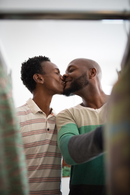 Close-up de casal gay africano se divertindo com beijos. dois homens sorridentes felizes em pé na loja de roupas escolhendo camisas em cabides se beijando. relações de casais do mesmo sexo, tempo livre para o conceito de compras