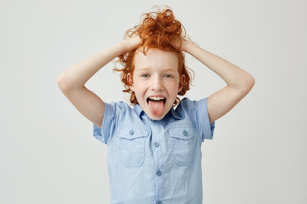 Close-up de criança bonita com cabelos cacheados laranja e sardas segurando o cabelo com as mãos, mostrando a língua, fazendo a expressão do rosto bobo