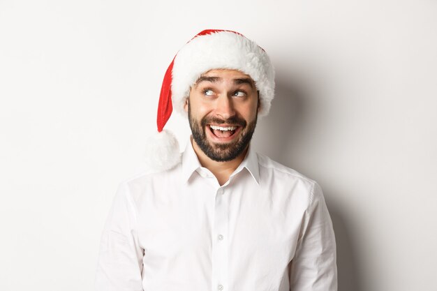 Close-up de feliz homem barbudo com chapéu de Papai Noel, celebrando o Natal e ano novo e olhando para o canto superior esquerdo, imaginando algo. fundo branco.