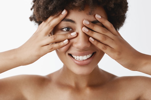 Foto grátis close-up de glamour emotiva e linda modelo feminina nua de pele escura com cabelos cacheados cobrindo os olhos com as palmas das mãos e espiando por entre os dedos sorrindo alegremente esperando por uma surpresa ou um presente