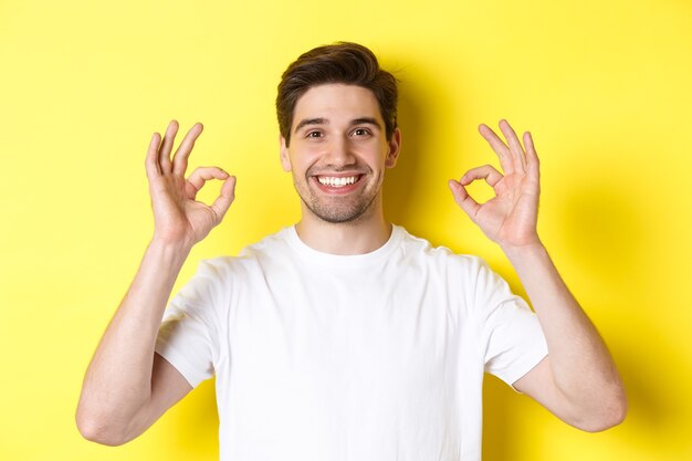 Close-up de jovem bonito mostrando sinal de tudo bem, aprovo e concordo, sorrindo satisfeito, em pé sobre fundo amarelo.