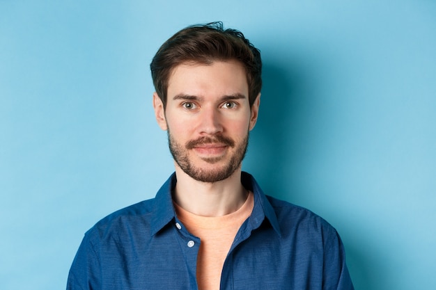 Close-up de jovem caucasiano com barba, sorrindo e olhando feliz para a câmera, de pé sobre fundo azul.