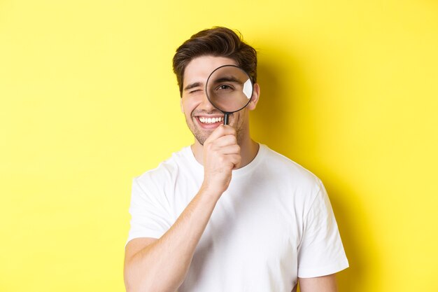 Close-up de jovem olhando pela lupa e sorrindo, procurando algo, em pé sobre um fundo amarelo.