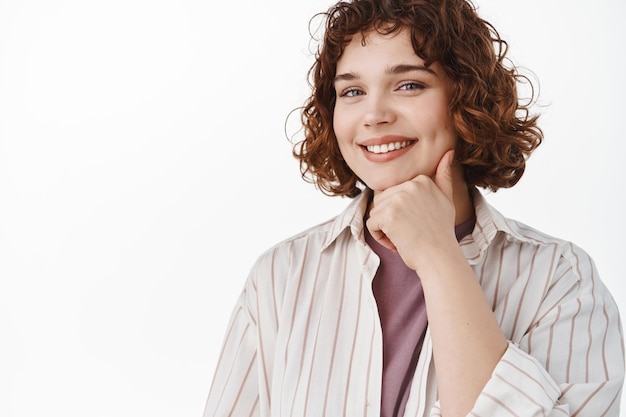 Close-up de linda garota sincera com cabelo curto encaracolado, sorrindo com dentes brancos e olhando pensativo para a câmera, verificando algo interessante, olhando para o anúncio, fundo branco