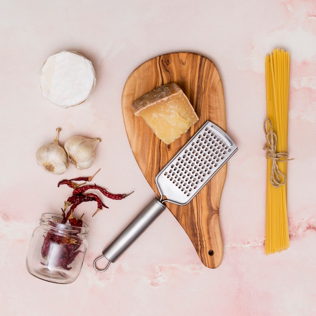 Close-up de macarrão cru; queijo; pimentão seco; utensílio de alho e cozinha em pano de fundo rosa