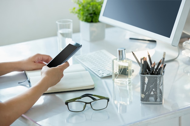 Close-up de mãos femininas, usando telefone inteligente enquanto trabalhava no computador no interior do escritório moderno