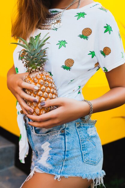 Foto grátis close-up de mãos segurando a pele bronzeada de abacaxi de uma mulher atraente de férias usando chapéu de palha descalça e shorts jeans com estampa de camiseta da moda de verão