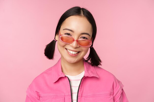 Close-up de menina coreana estilosa em óculos de sol sorrindo feliz posando contra fundo rosa Conceito de rosto de pessoas