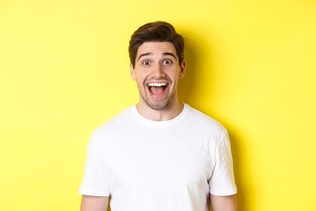 Close-up de um cara bonito surpreso, reagindo a uma ótima notícia, em pé sobre um fundo amarelo em uma camiseta branca.