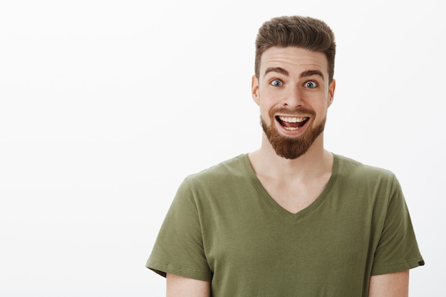 Close-up de um homem barbudo feliz, surpreso e animado, erguendo as sobrancelhas e arregalando os olhos, sorrindo amplamente ao receber uma oferta incrível e inesperada de toda a vida sobre uma parede branca