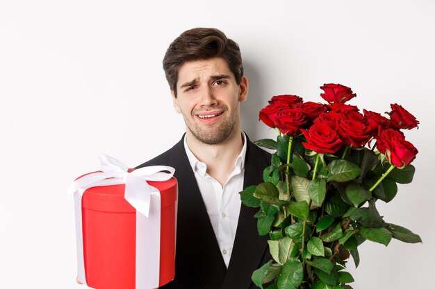 Close-up de um homem cético de terno, segurando um buquê de rosas vermelhas e um presente, relutante contra um fundo branco.