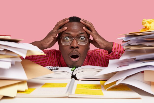 Close up de um homem negro afro-americano chocado e estupefato com as duas mãos na cabeça, olhando uma pilha de livros e papéis, tendo uma tarefa difícil