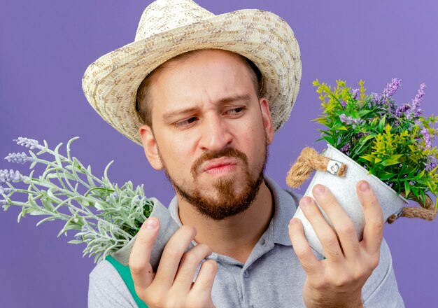 Close-up de um jovem bonito e confuso jardineiro eslavo de uniforme e chapéu segurando e olhando vasos de flores isolados na parede roxa