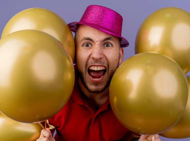 Close-up de um jovem bonito e furioso com um chapéu de festa, olhando para a frente, segurando balões isolados na parede roxa