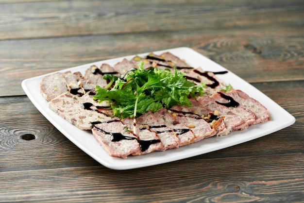 Close-up de um prato quadrado branco servido com fatias de carne recheada, decorado com folhas verdes e molho de soja na mesa de madeira.