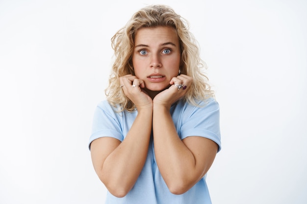 Close-up de uma fêmea fofa insegura e boba com medo de cabelos loiros e olhos azuis cerrando os dentes tremendo de medo, segurando as mãos na linha do queixo, preocupação sendo perturbada e confusa, expressando nervosismo