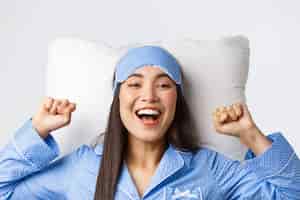 Foto grátis close-up de uma garota asiática entusiasmada de pijama azul e máscara de dormir, esticando as mãos encantada após uma boa noite de sono, máscara de decolagem pela manhã, deitada na cama no travesseiro e sorrindo feliz